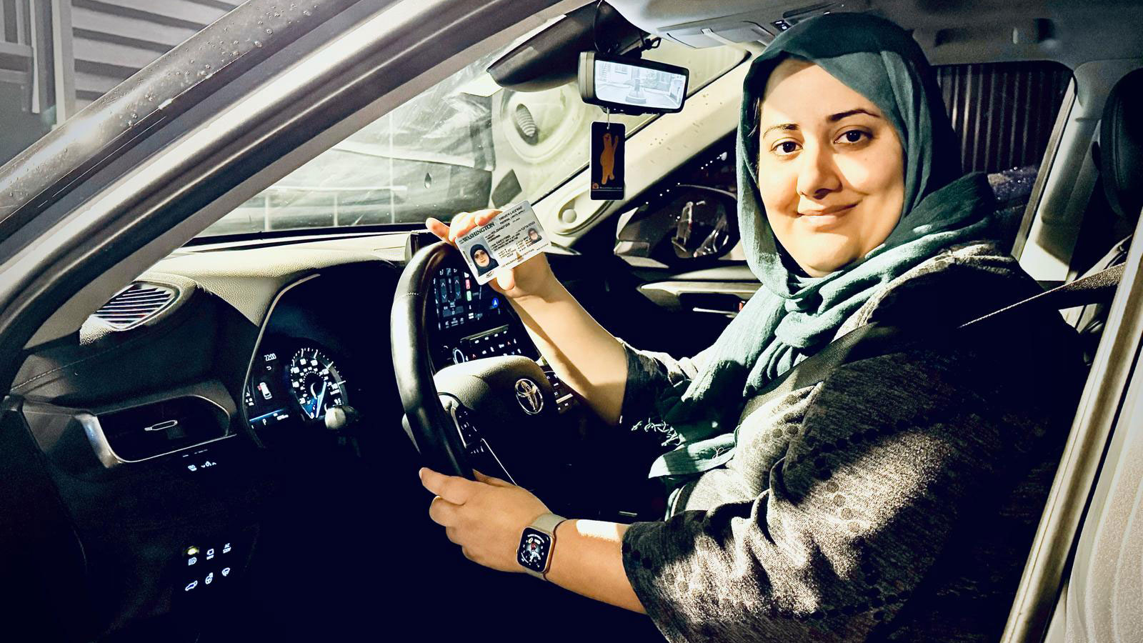 Woman wearing a hijab sitting in the driver's seat. She looks slightly over her shoulder smiling at the camera. She holds a driver's license in her hand.